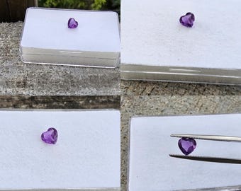 Könnte beinhalten: Ein herzförmiger, facettierter Amethyst-Edelstein. Der lila Edelstein wird in einer transparenten Kunststoffbox mit weißem Innenraum präsentiert. Der Edelstein wird auch von einer Pinzette gehalten. Der Hintergrund ist eine natürliche Umgebung im Freien.