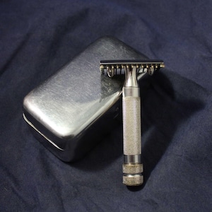 May include: A silver safety razor with a textured handle and a matching metal case. The razor is designed for shaving and has a classic, vintage aesthetic. The case is rectangular and has rounded edges.