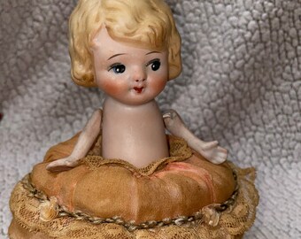 May include: A vintage porcelain doll with blonde hair and blue eyes, sitting on a round, brown, fabric-covered cushion with lace trim.