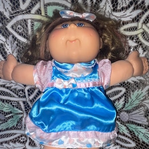 May include: A vintage Cabbage Patch Kids doll with a soft, plush body. The doll has blue eyes, rosy cheeks, and brown hair. It wears a blue satin dress with pink trim and a matching bow. The doll's dress has a white lace collar.