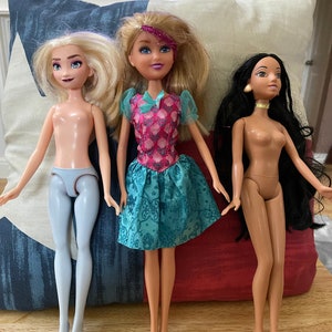 K&ouml;nnte beinhalten: Drei Puppen mit langen Haaren. Die Puppe links hat blonde Haare und tr&auml;gt ein hellblaues Kleid. Die Puppe in der Mitte hat blonde Haare und tr&auml;gt ein pinkfarbenes und t&uuml;rkisfarbenes Kleid. Die Puppe rechts hat lange schwarze Haare und tr&auml;gt ein braunes Kleid.