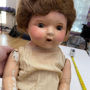 May include: Antique porcelain doll with brown hair, wearing a white dress and a white skirt. The doll's face has cracks and the paint is chipped. The doll's arms are cracked and the paint is peeling.