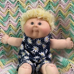 May include: A vintage Cabbage Patch Kid doll with blonde yarn hair and brown eyes. The doll wears a navy blue romper with a white daisy pattern. The doll is sitting on a pastel-colored, chevron-patterned blanket.
