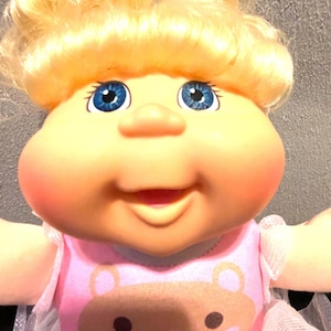 May include: A Cabbage Patch Kids doll with blonde curly hair and blue eyes. The doll is wearing a pink dress with a bear graphic. The doll's cheeks are rosy, and it has a smiling expression.