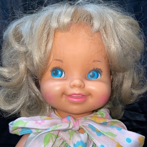 May include: A vintage doll with blonde hair, blue eyes, and a pink and white floral dress. The doll has a smiling expression and rosy cheeks.