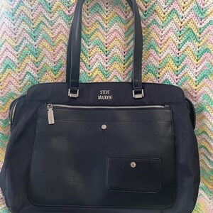 May include: Black tote bag with two handles and a shoulder strap. The bag has a front pocket and a zippered top. The brand name "STEVE MADDEN" is visible on the front of the bag. The bag is made of a nylon material.