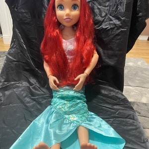 May include: A large doll of a mermaid princess with red hair, blue eyes, and a purple tiara. The doll wears a pink top and a turquoise mermaid tail. The doll is approximately 30 inches tall.