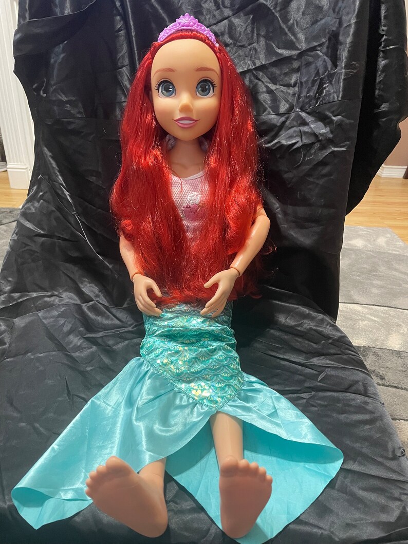 May include: A large doll of a mermaid princess with red hair, blue eyes, and a purple tiara. The doll wears a pink top and a turquoise mermaid tail. The doll is approximately 30 inches tall.