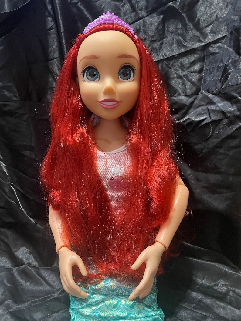 May include: A large doll of a character with red hair, blue eyes, and a purple tiara. The doll wears a pink top with a cupcake design and a teal skirt with a scale pattern. The doll's arms are bent at the elbows.