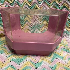 May include: A pink plastic display case with a clear plastic top. The case has a hexagonal shape and is sitting on a colorful, patterned blanket. The case appears to be vintage.