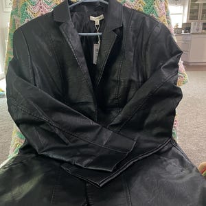 May include: A black faux leather blazer with a notched collar and long sleeves, hanging on a blue hanger. The jacket has a tailored fit and a slightly flared hem. A price tag hangs from the jacket.