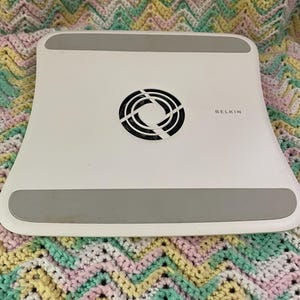 May include: A white Belkin laptop cooling pad with a curved rectangular shape. The device features a central circular black fan vent and two horizontal gray non-slip rubber strips positioned at the top and bottom edges of the white plastic surface.