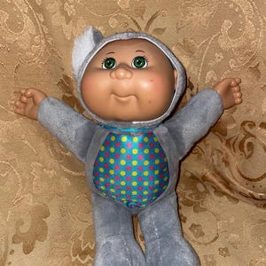 May include: A gray plush Cabbage Patch Kid doll wearing a gray koala costume with a blue, pink, and yellow polka dot pattern on the chest.