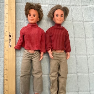 May include: Two vintage dolls with brown hair, wearing red ribbed sweaters and tan pants. One doll has bare feet, the other has brown shoes. A wooden ruler is on the left side of the dolls.