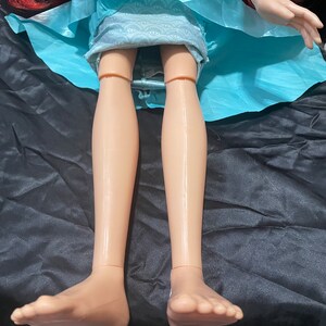 May include: A doll with red hair, wearing a light blue dress. The doll's legs and feet are visible, with a tan skin tone. The doll is lying on a black surface.