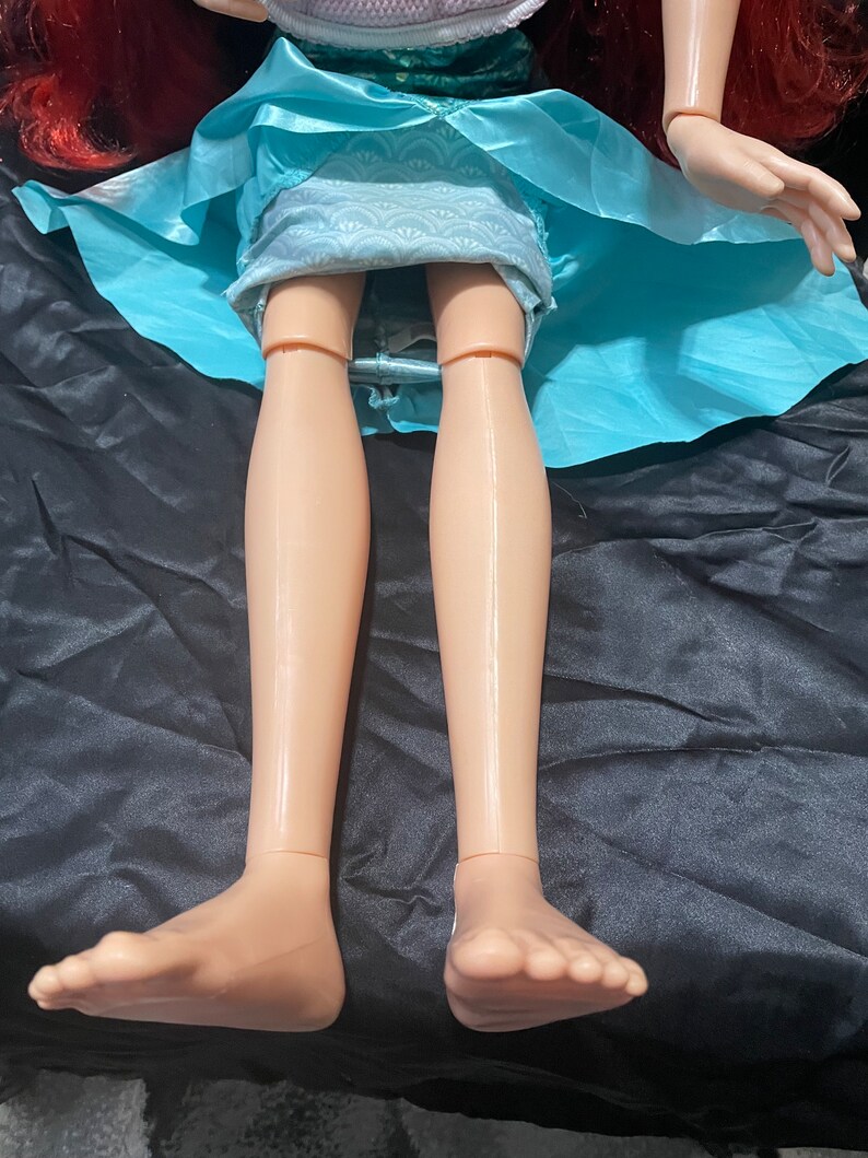 May include: A doll with red hair, wearing a light blue dress. The doll's legs and feet are visible, with a tan skin tone. The doll is lying on a black surface.