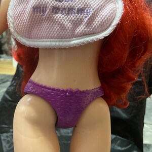 May include: A doll with red hair, wearing a pink bib with the text "I love my mommy" and a purple bottom. The bib has a white trim and a pink embroidered design of a flower.