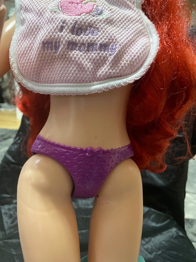 May include: A doll with red hair, wearing a pink bib with the text "I love my mommy" and a purple bottom. The bib has a white trim and a pink embroidered design of a flower.