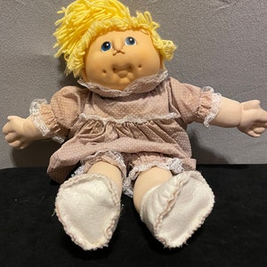 May include: A vintage Cabbage Patch Kid doll with blonde hair, blue eyes, and a pink and white polka dot dress. The doll is sitting with its legs crossed and wearing white socks.
