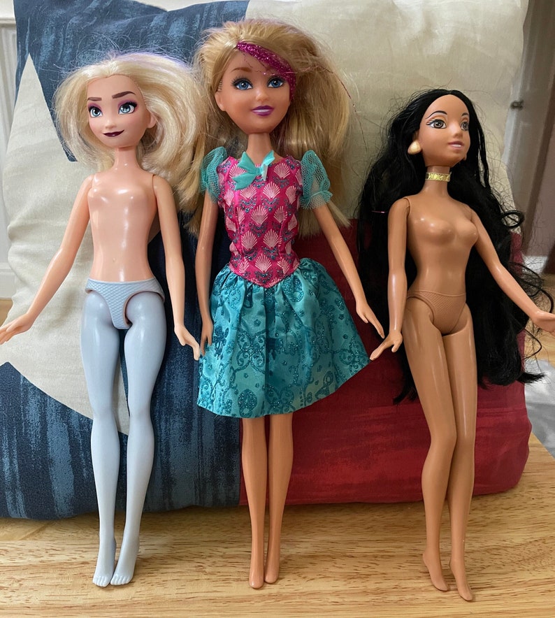 K&ouml;nnte beinhalten: Drei Puppen mit langen Haaren. Die Puppe links hat blonde Haare und tr&auml;gt ein hellblaues Kleid. Die Puppe in der Mitte hat blonde Haare und tr&auml;gt ein pinkfarbenes und t&uuml;rkisfarbenes Kleid. Die Puppe rechts hat lange schwarze Haare und tr&auml;gt ein braunes Kleid.