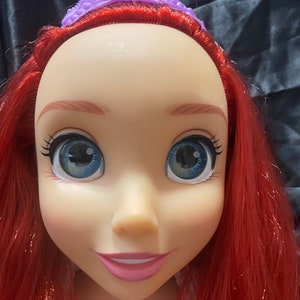 May include: A doll's head with red hair, blue eyes, and a purple tiara. The doll has a fair complexion, pink lips, and long eyelashes. The hair is styled in long waves. The tiara is a detailed, ornate design.