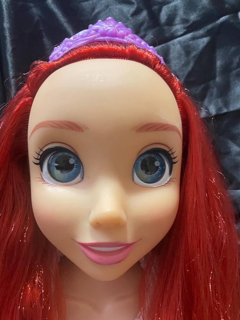 May include: A doll's head with red hair, blue eyes, and a purple tiara. The doll has a fair complexion, pink lips, and long eyelashes. The hair is styled in long waves. The tiara is a detailed, ornate design.