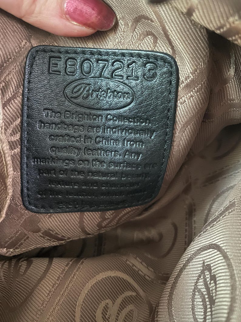 Może przedstawiać: Zbliżenie czarnej sk&oacute;rzanej metki z torebki Brighton. Metka ma numer "E807213" i logo Brighton. Tekst na metce brzmi "The Brighton Collection handbags are individually crafted in China from quality leathers."
