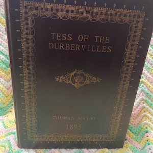 May include: A vintage hardcover book titled "Tess of the d'Urbervilles" by Thomas Hardy, published in 1895. The book has a dark brown cover with gold decorative accents and lettering. The book is resting on a colorful, patterned surface.