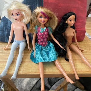 K&ouml;nnte beinhalten: Drei Puppen sitzen auf einem Holztisch. Die Puppe links hat blonde Haare und tr&auml;gt ein hellblaues Kleid. Die Puppe in der Mitte hat blonde Haare und tr&auml;gt ein blaues und rosa Kleid. Die Puppe rechts hat lange schwarze Haare und tr&auml;gt ein braunes Kleid.