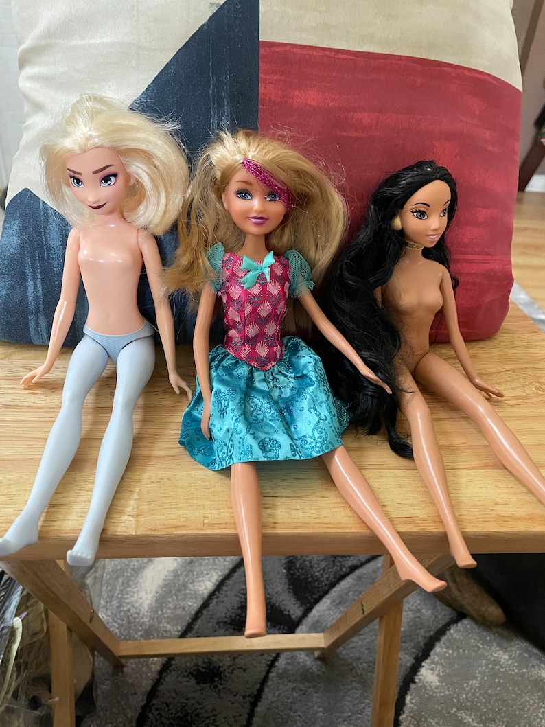 K&ouml;nnte beinhalten: Drei Puppen sitzen auf einem Holztisch. Die Puppe links hat blonde Haare und tr&auml;gt ein hellblaues Kleid. Die Puppe in der Mitte hat blonde Haare und tr&auml;gt ein blaues und rosa Kleid. Die Puppe rechts hat lange schwarze Haare und tr&auml;gt ein braunes Kleid.