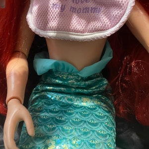 May include: Ariel doll with red hair and a teal mermaid tail. The doll wears a pink bib with the text "I love my mommy". The doll's arm is visible.