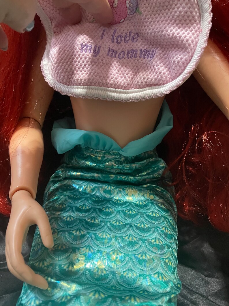 May include: Ariel doll with red hair and a teal mermaid tail. The doll wears a pink bib with the text "I love my mommy". The doll's arm is visible.