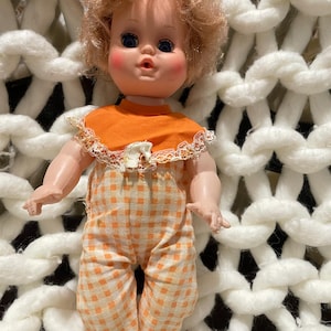 May include: A vintage doll with blue eyes, rosy cheeks, and curly blonde hair. The doll wears an orange bib with lace trim and orange and white checkered pants. The doll is posed against a white, chunky knit background.