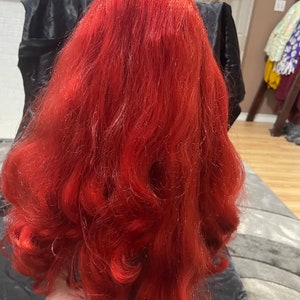 May include: A doll with long, vibrant red hair styled in loose curls. A purple tiara sits atop the head. The doll's hair is the focal point, with a rich, saturated color and a flowing texture.
