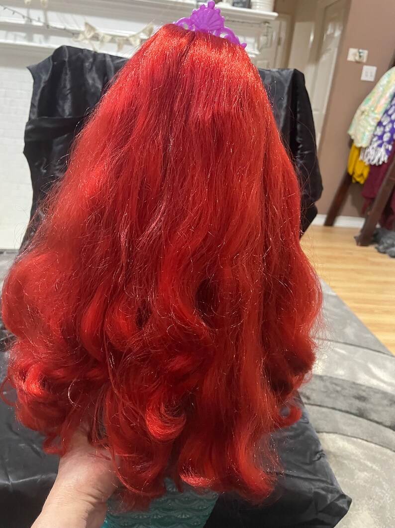 May include: A doll with long, vibrant red hair styled in loose curls. A purple tiara sits atop the head. The doll's hair is the focal point, with a rich, saturated color and a flowing texture.
