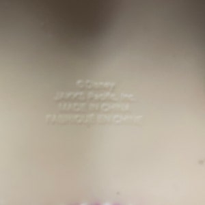 May include: A close-up of a light beige surface with the text "&copy;Disney", "JAY@S Pacific Inc.", "MADE IN CHINA", and "FABRIQUE EN CHINE". A purple scalloped edge is visible at the bottom.