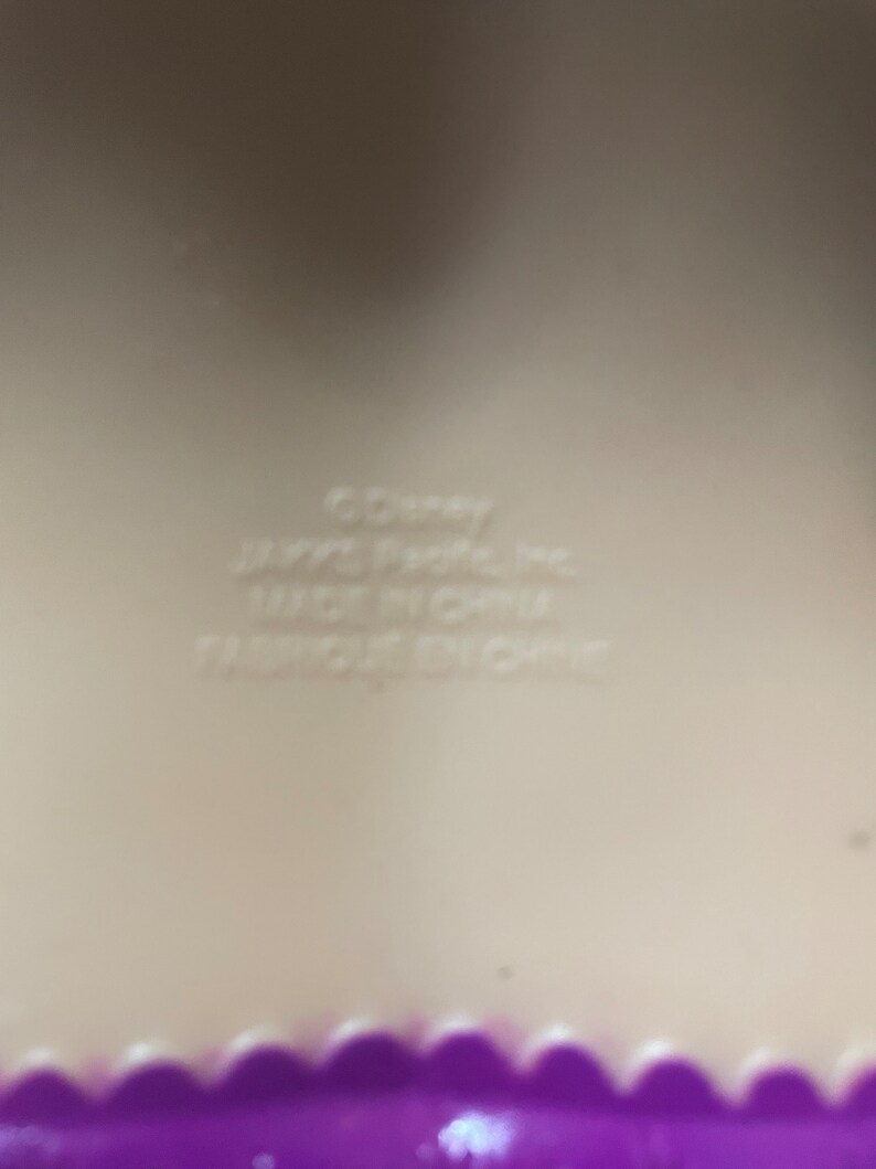 May include: A close-up of a light beige surface with the text "&copy;Disney", "JAY@S Pacific Inc.", "MADE IN CHINA", and "FABRIQUE EN CHINE". A purple scalloped edge is visible at the bottom.