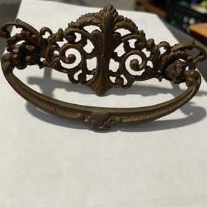 May include: Ornate bronze-colored drawer pull with a curved handle and decorative floral and scrollwork details. The central design features a stylized leaf motif, and the ends of the handle have small, detailed floral accents. This vintage-style hardware is suitable for furniture restoration.