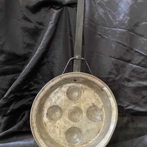 May include: A vintage silver-colored metal pan with a black metal handle. The pan has five round indentations in the bottom, likely for making individual cakes or pastries.