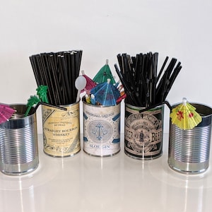 Op de afbeelding: Vier lege blikjes met kleurrijke papieren cocktailparasols erop. De blikjes zijn gelabeld met verschillende alcoholmerken, waaronder "Straight Bourbon Whiskey", "Sloe Gin" en "Continental Brewing Co. Lager Beer".