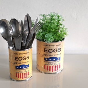 Può includere: Due barattoli di metallo in stile vintage con un design a bandiera americana rosso, bianco e blu. Entrambi i barattoli hanno stampate le parole "Pure Dried Whole Eggs" e "Net Weight 5 Ounces". Un barattolo contiene un set di posate, mentre l'altro contiene una piccola pianta in vaso.