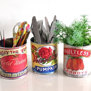 Może przedstawiać: Trzy metalowe puszki w stylu vintage, przerobione na pojemniki. Puszka po lewej stronie jest czerwona z niebieskimi i złotymi obw&oacute;dkami i napisami "Defy the World Cold Packed Tomatoes". Puszka w środku jest ż&oacute;łta z niebieskimi i czerwonymi obw&oacute;dkami i napisami "Velvet Rose Pumpkin". Puszka po prawej stronie jest biała z czerwonymi i niebieskimi obw&oacute;dkami i napisami "Faultless Brand Carrots".