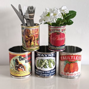 May include: Five vintage-style metal cans with different labels. The cans are used as containers for utensils, flowers, and other items. The labels feature images of a lion, tomatoes, butterflies, green beans, and carrots. The labels also include text such as "Lion Brand", "Defy the World", "Butterfly Brand", "Wayne", and "Faultless Brand".