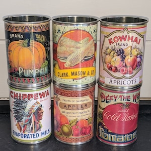 Pode incluir: Seis latas de metal vintage com rótulos coloridos que representam várias frutas e vegetais. Os rótulos incluem "Golden Pumpkin", "Clark, Mason & Co.", "Apricots", "Chippewa", "Apples" e "Defy the World Cold Sacked Tomatoes".