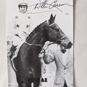 May include: A black and white photograph of a jockey on a horse with a man holding the reins. The jockey is wearing a white uniform with stars on the shoulders. The photo is signed "Best Wishes, Willie Carson".