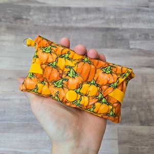 May include: A small, orange fabric wallet with a pumpkin pattern. The wallet has a yellow button closure and a yellow strap. The pumpkin pattern features orange pumpkins with green stems. A small metal ring is attached to the top.