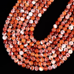 May include: A collection of round, polished beads arranged in a curved formation. Each bead features a marbled pattern with deep red, orange, and white tones, creating a cracked or webbed appearance across the smooth, spherical surfaces.