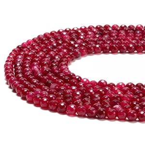 May include: Close-up of strands of faceted, deep red gemstone beads. The beads are round and have a polished, reflective surface. The beads are strung together, creating a flowing, organic shape. The color is a rich, saturated red.