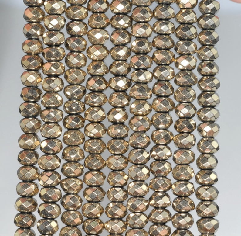 May include: Close-up of strands of faceted, golden-colored pyrite beads. The beads have a metallic sheen and are strung together, creating a textured surface. The beads are oval-shaped with multiple facets, reflecting light. Ideal for jewelry making.