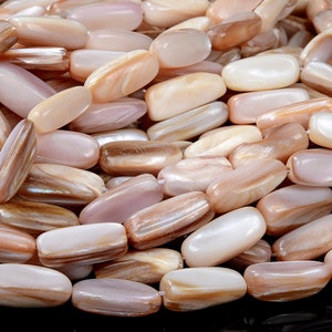 Puede incluir: Primer plano de un montón de cuentas ovaladas, iridiscentes, de nácar. Las cuentas son de una mezcla de colores rosa claro, blanco y marrón.